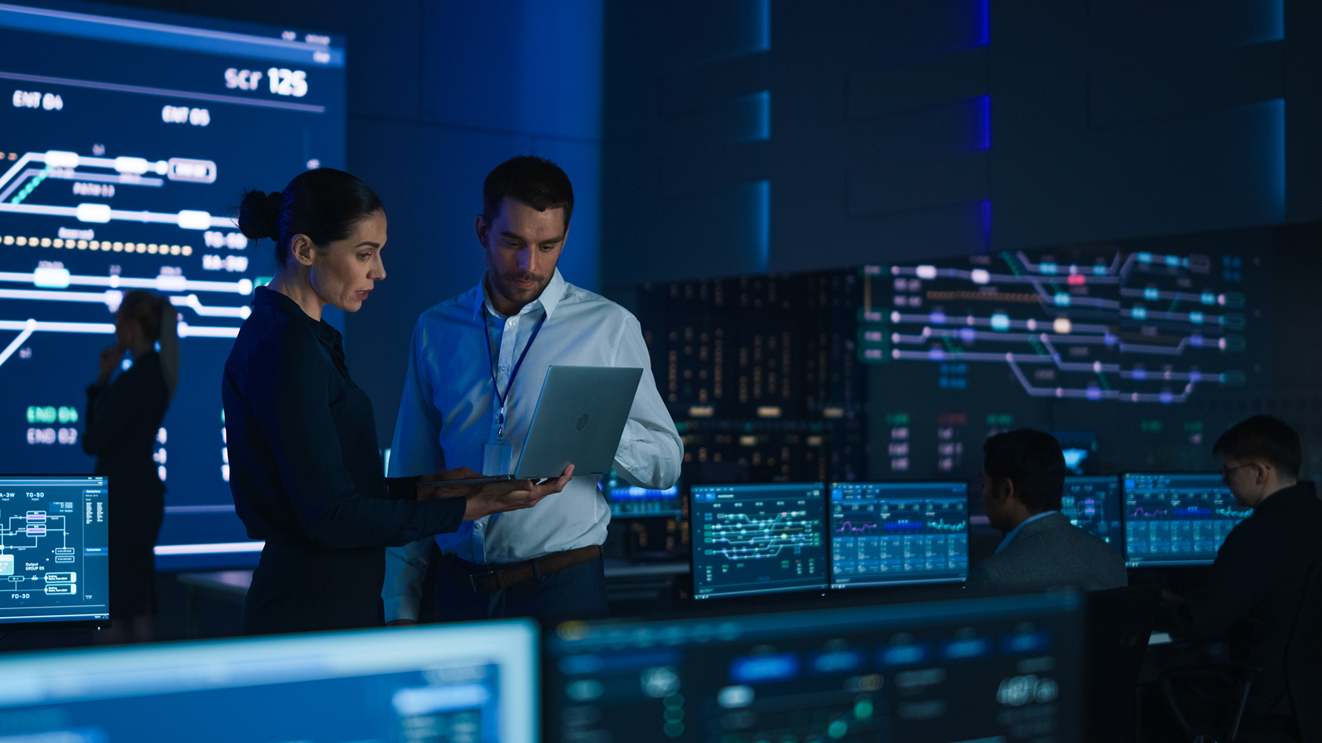 project leader and chief executive discuss data shown on a laptop. in background big screen showing infographics of infrastructure, charts, system analysis. telecommunications control room